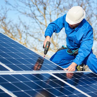 Männlicher Ingenieur in blauem Anzug und Schutzhelm bei der Installation von Photovoltaikmodulen mit einem Schraubenzieher. Professioneller Elektriker bei der Montage von Solarmodulen auf dem Dach. Ökologisches Konzept der alternativen Energie.