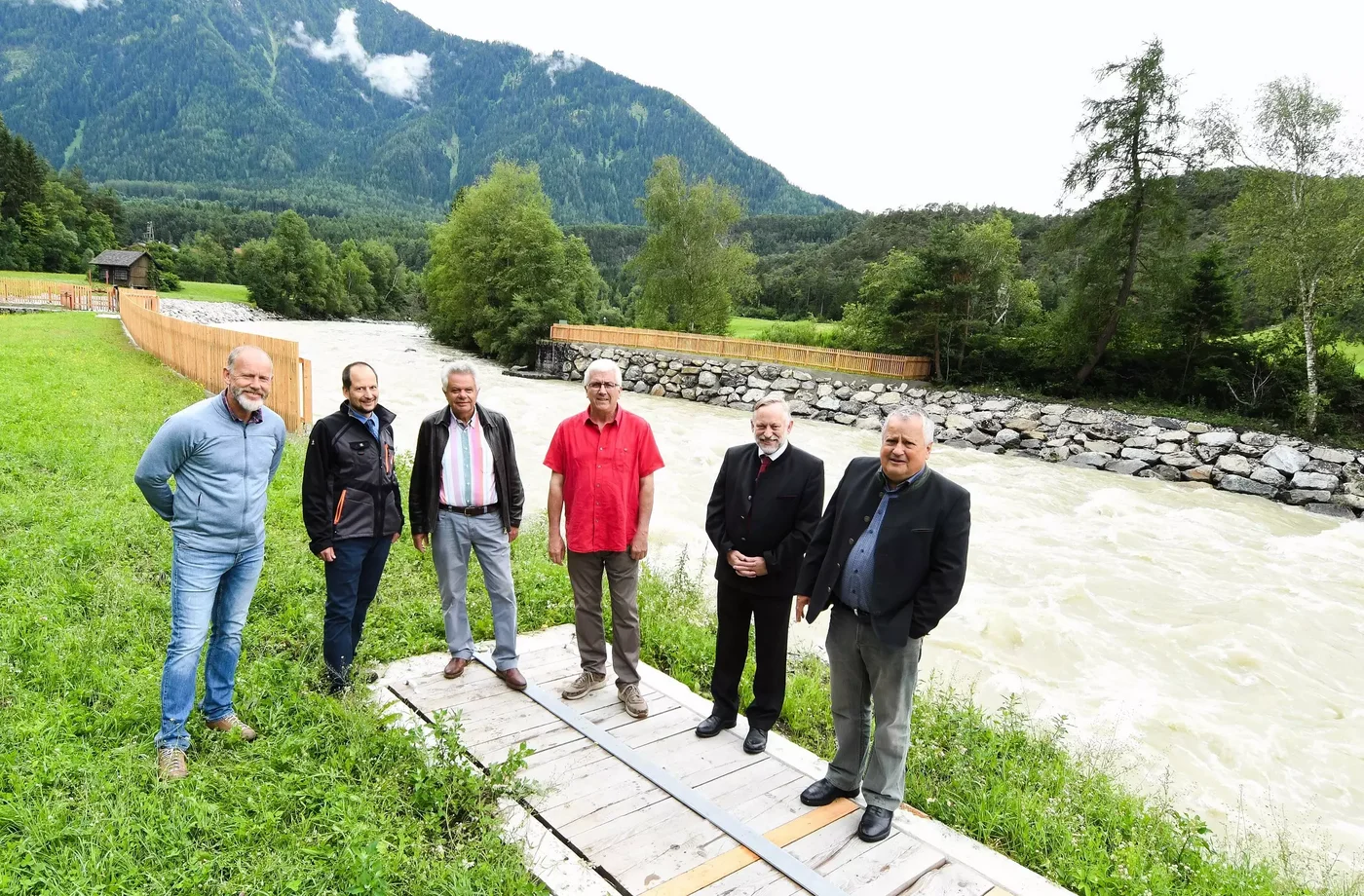 Sechs Männer stehen lachend vor einem Bach nebeneinander.