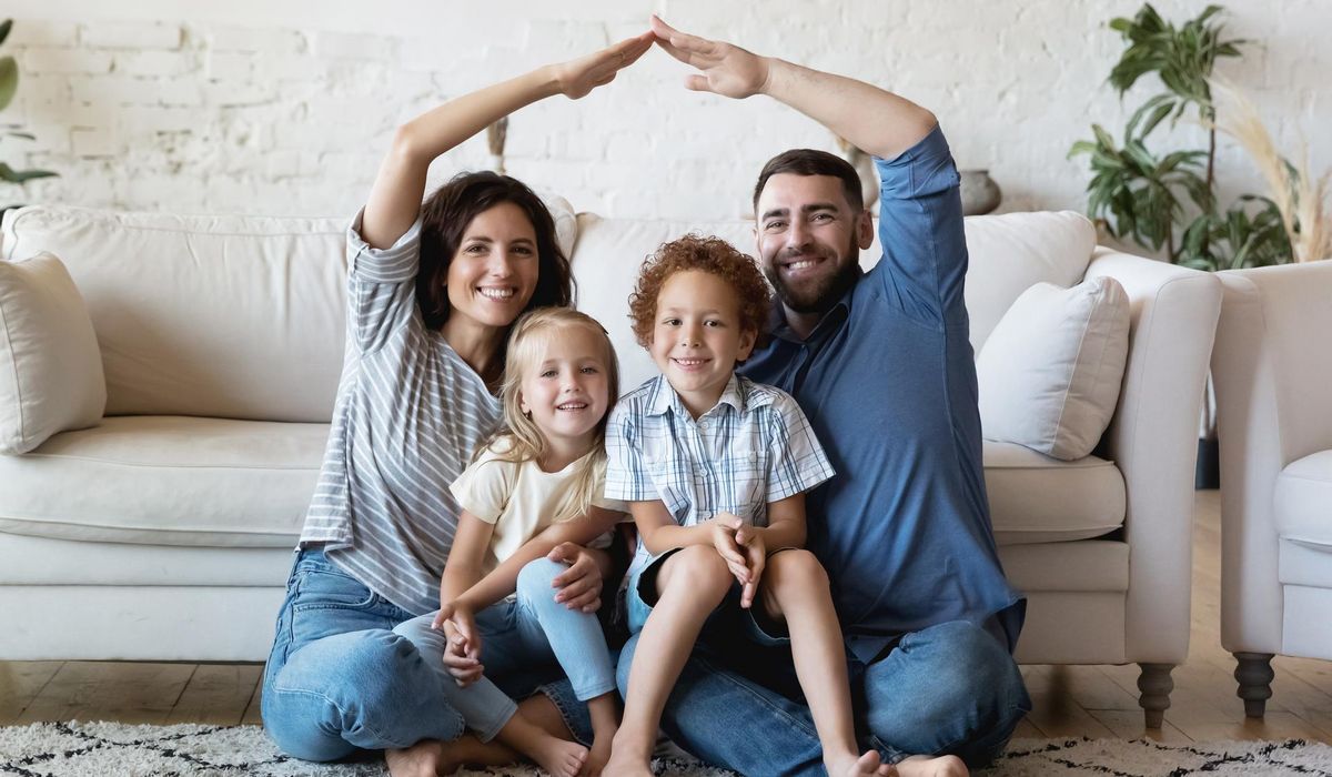 Eine Familie sitzt vor dem Sofa auf einem Teppich und lächeln. Die zwei Kinder sitzen auf dem Schoß ihrer Eltern. Die Eltern bilden mit den Händen ein Dach.