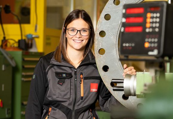 Eine Frau steht lachend in ihrer Arbeitskleidung neben einer Maschine.