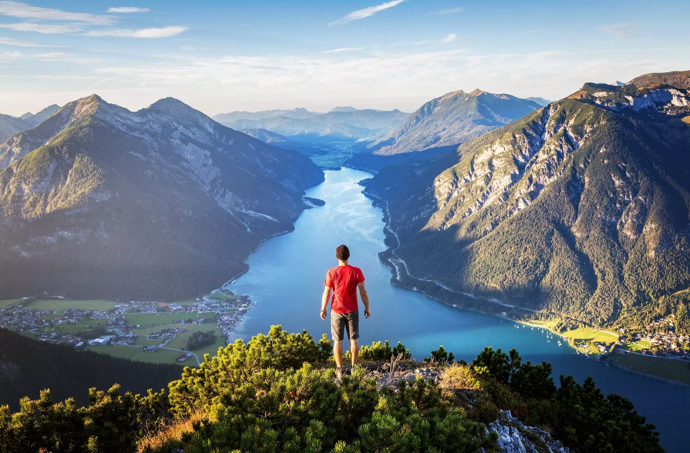 Ein Mann steht auf einem Berg und schaut hinunter. Dort sieht man einen großen See und Berge sowie die Häuser im Tal.