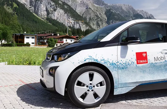 TIWAG - E-Auto parkt vor einer Ladestation. Im Hintergrund sind Berge, Häuser und grüne Felder.
